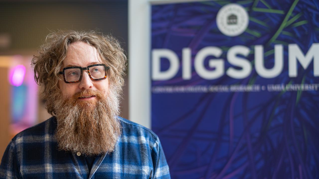 En man med skägg och glasögon står framför en skylt.