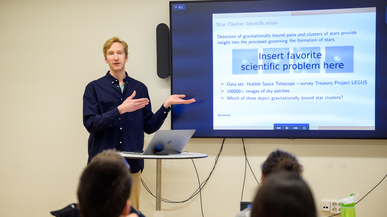 Jakob Hultgren speaks facing a seated crowd , to the side of him is a screen showing text: Insert favorite scientific problem here.