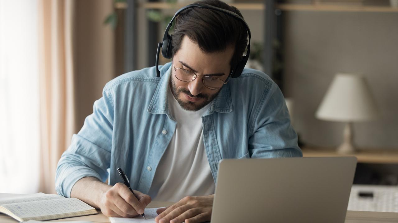 En man sitter vid ett bord med laptop och headset, böcker och antecknar i ett block.
