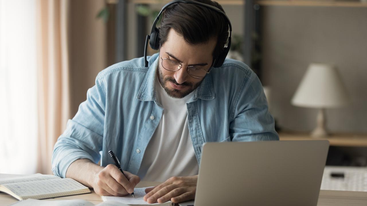 En man sitter vid ett bord med laptop och headset, böcker och antecknar i ett block.