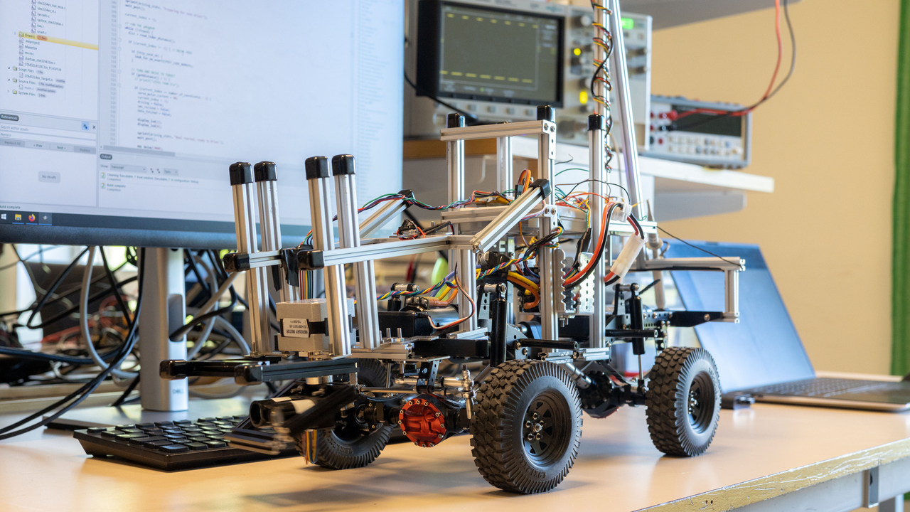 Ett autonomt fordon står på ett skrivbord. Det är en robot. I bakgrunden ser man dator-utrustning och program på bland annat skärn.