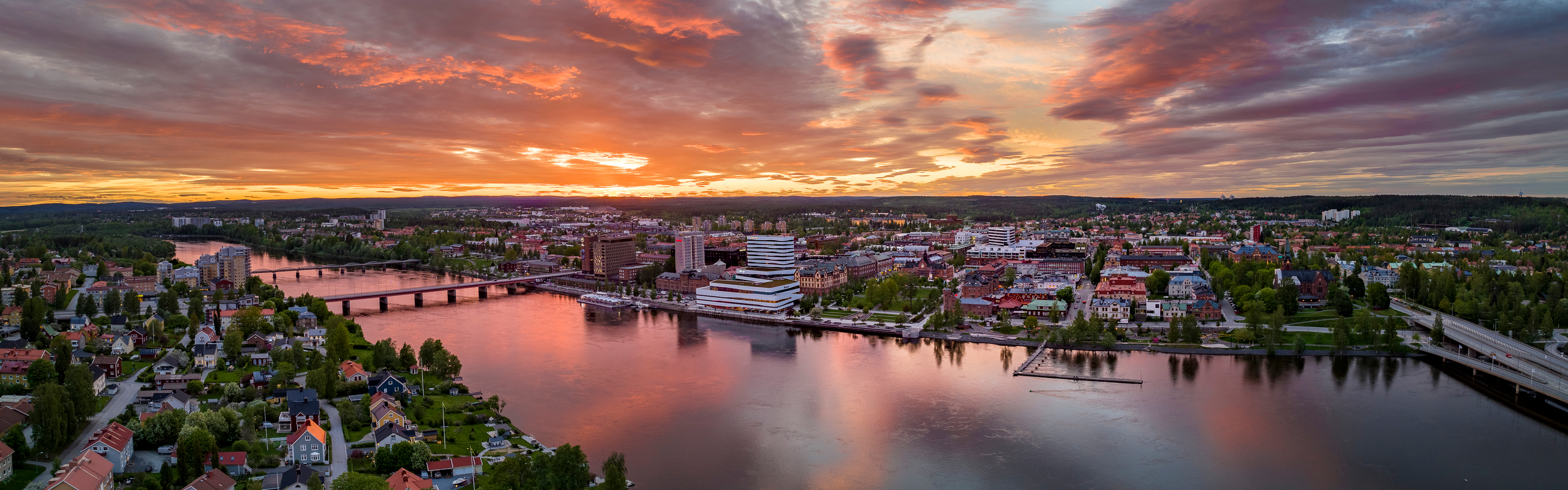 Vy tagen med drönare från södra sidan av Umeåälven mot Umeås centrum med kulturhuset Väven i fokus. Himlen är röd-orange i solnedgången.