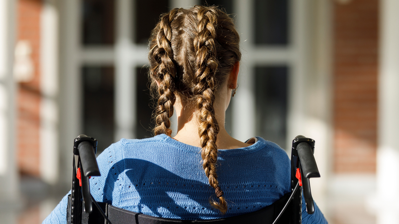 Flicka med flätor sitter i rullstol framför byggnad.
