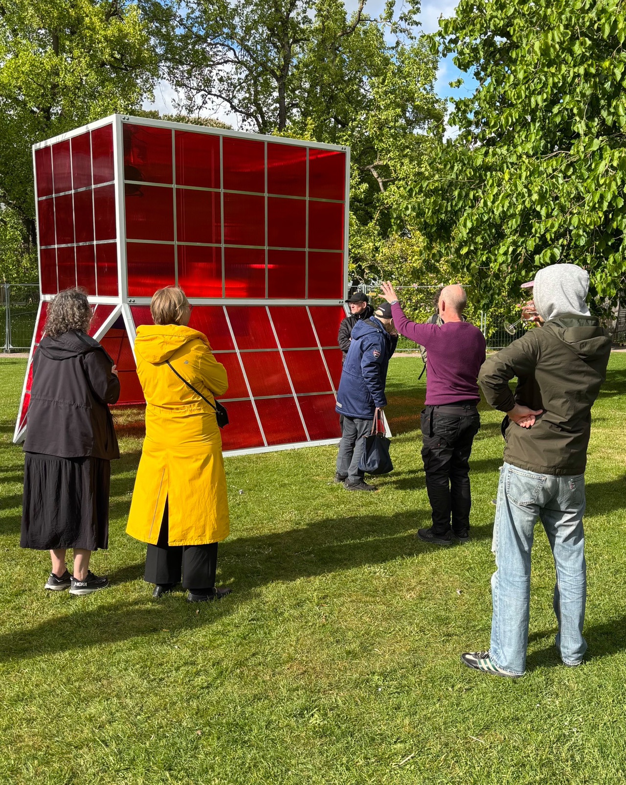 Människor står på en grön gräsmatta och beundrar en stor skulptur som liknar ett växthus i  rött glas.