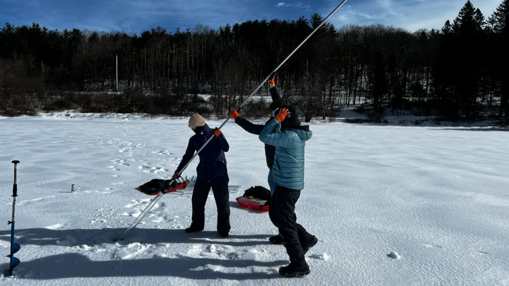 Tre människor för en lång borr ned i ett hål i isen på en sjö.