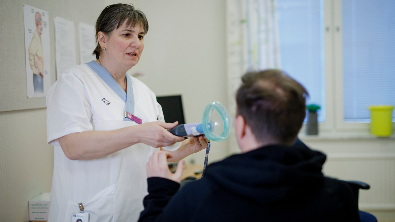 In an examination room research nurse Elisabeth Müller Granberg extends a spirometer to the ALS patient, who is sitting with his back to the camera.