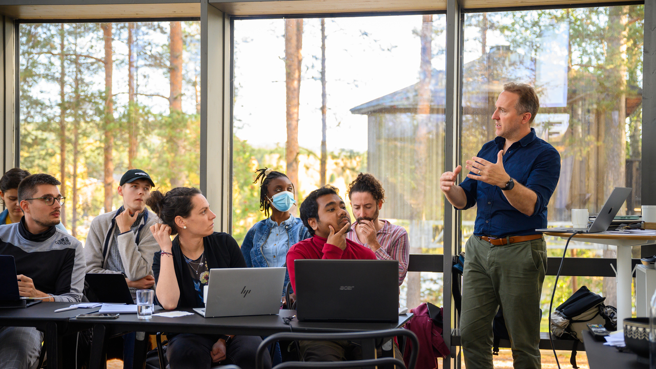 A group of students sitting at tables with laptops open listen to a man standing and speaking while gesturing with his arms. They are in a glass enclosed room, trees and another wooden house are visible outside. 