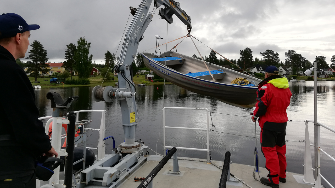 Två personer står på en större båt och lyfter en mindre båt med lyftkran