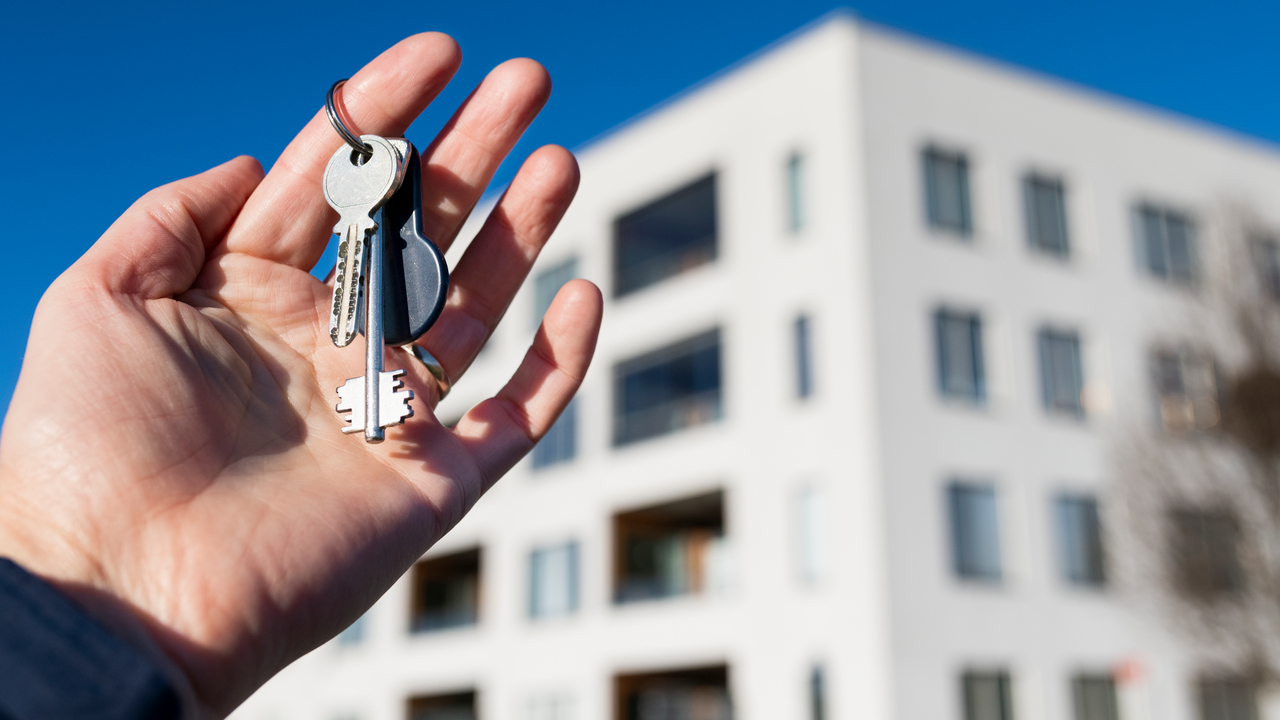 Hand med nycklar till en lägenhet. Lägenhetshus i bakgrunden.