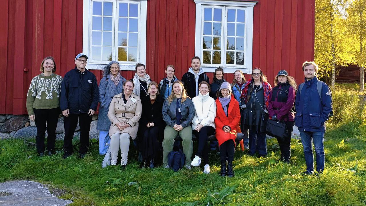 En grupp människor står och ler mot kameran. I bakgrunden har de ett rött trähus med stora vita fönster.