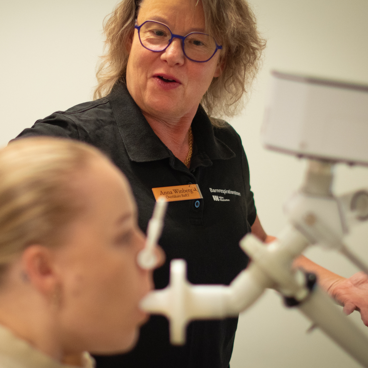 En kvinna blåser i en spirometer medan en kvinna i svart instruerar henne.