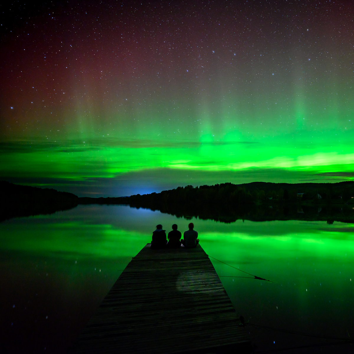 A group of people sitting on a dock watching the aurora reflecting in a river.