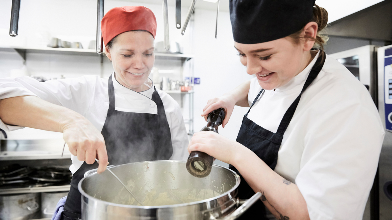 Två glada personer i förkläden och kockmössor runt en stor gryta.