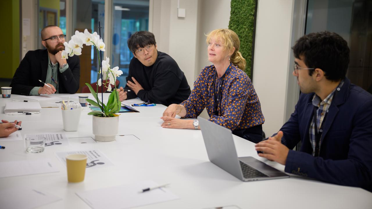 Four people sit at a table. The woman who is third from the left is speaking while the others, three men, listen.