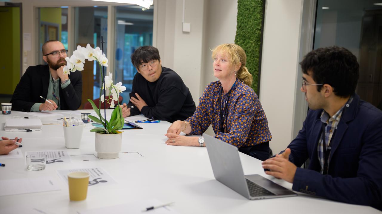 Four people sit at a table. The woman who is third from the left is speaking while the others, three men, listen.