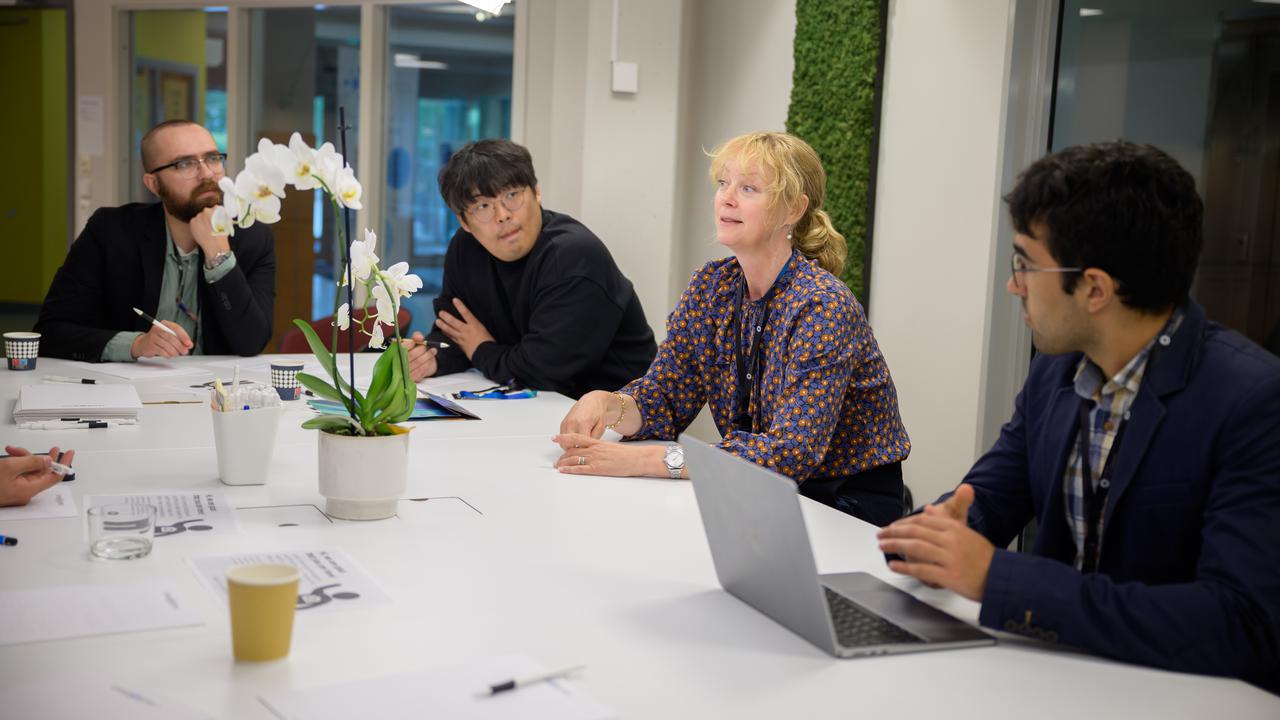 Four people sit at a table. The woman who is third from the left is speaking while the others, three men, listen.