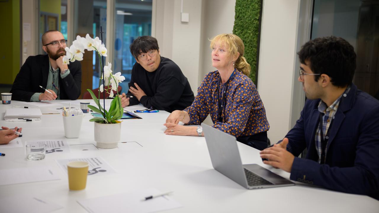 Four people sit at a table. The woman who is third from the left is speaking while the others, three men, listen.