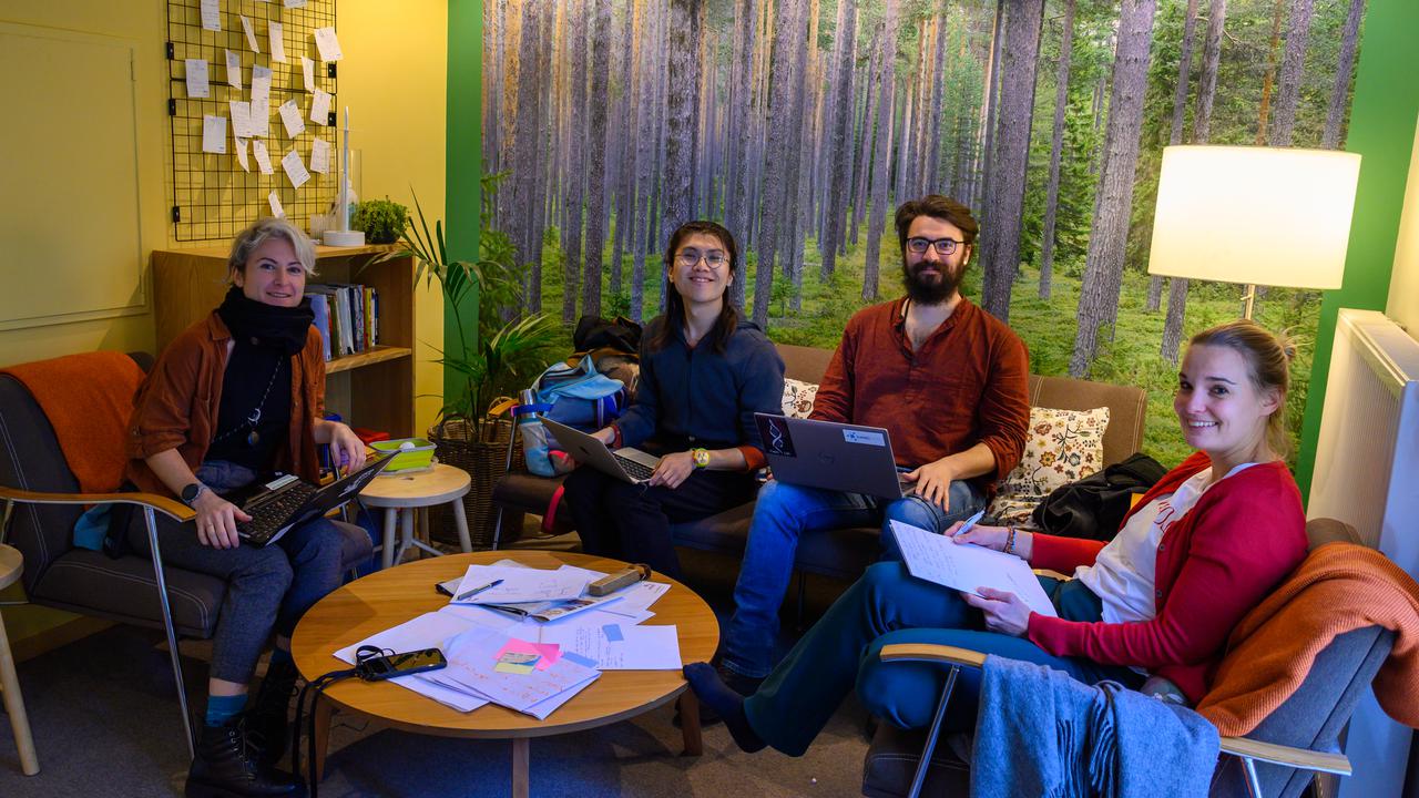 Four people smile at the camera from their seated positions at a table working together