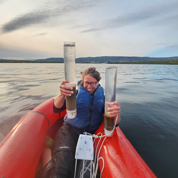 Václava Hazuková samlar in sedimentprovet från en arktisk sjö. Hon sitter i en liten röd gummibåt och håller upp två stora cylindriska behållare med vatten och sediment. 
