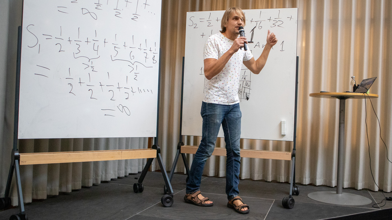 Niklas Lundström, universitetslektor, på scenen i MIT-huset, bakom honom whiteboardtavlor med mattetal