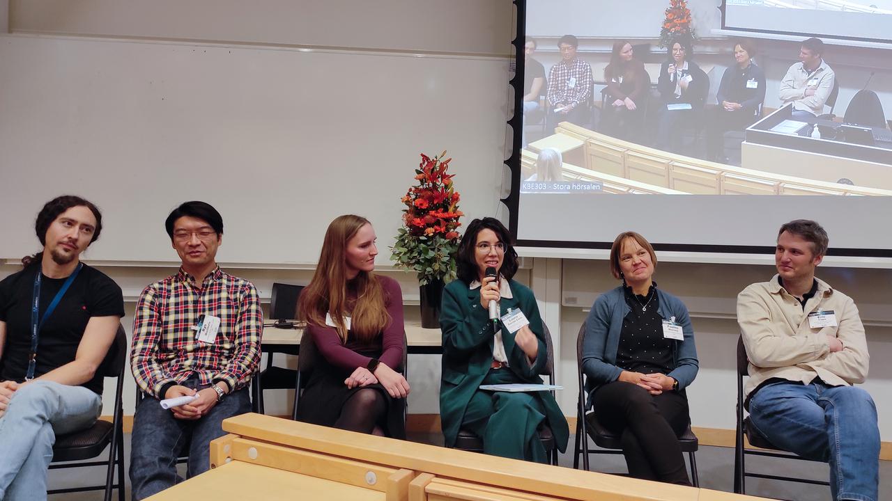 A group of six panel members sits in front of a projector screen, one person speaks into the microphone