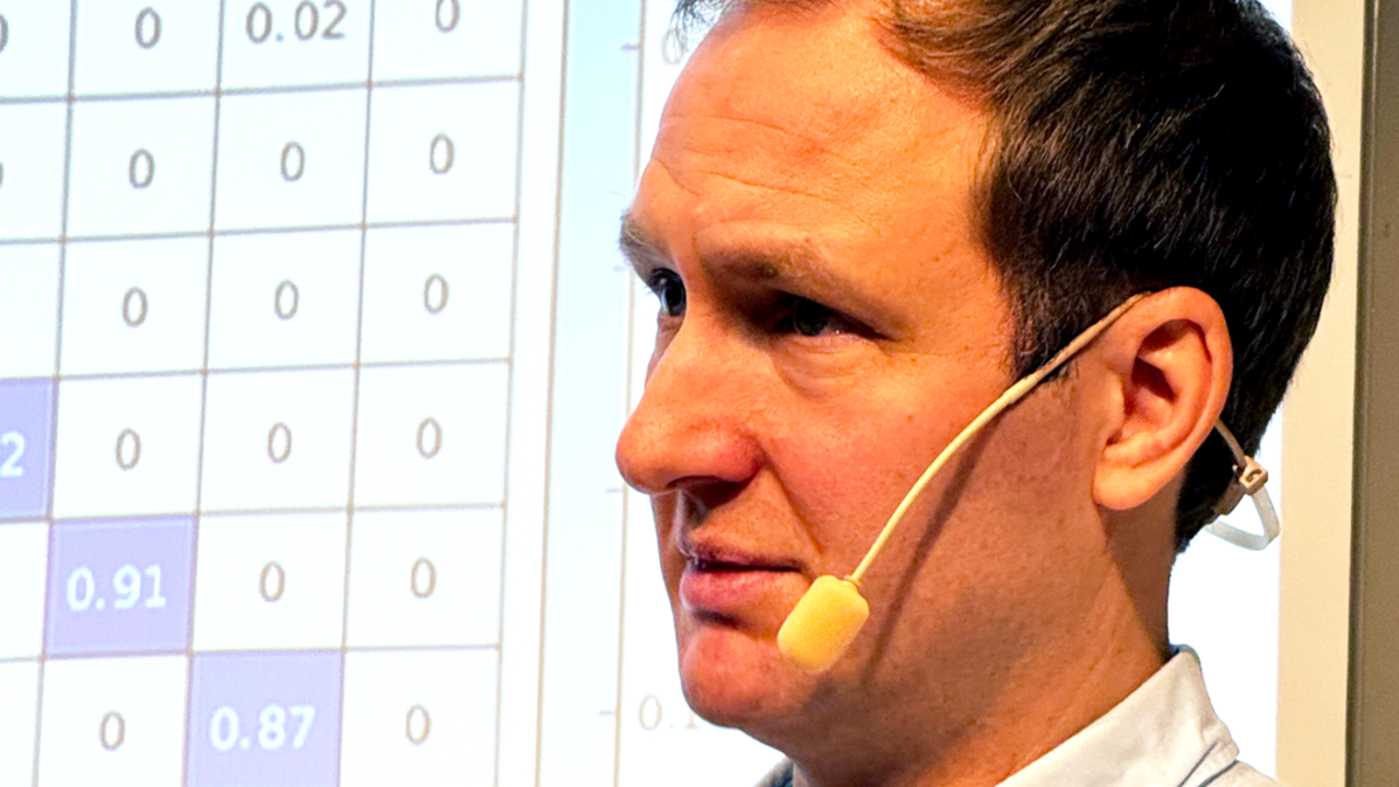 Man framför filmduk som visar en matematisk matris. Han bär ljusblå skjorta och han har en mikrofonmygga kring örat.