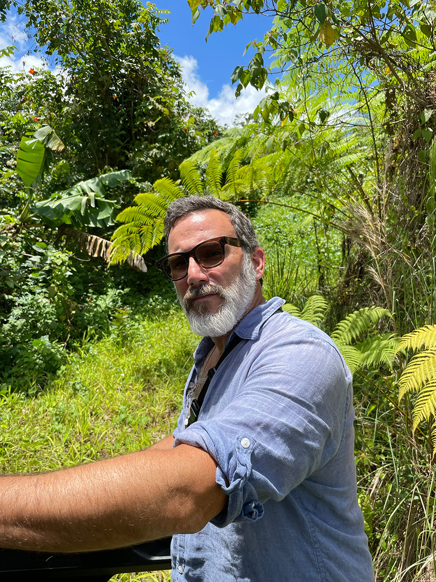 Luis Berrios-Negrón står i förgrunden med en blå skjorta och solglasögon på sig. Han omges av grön regnskog och blå himmel.