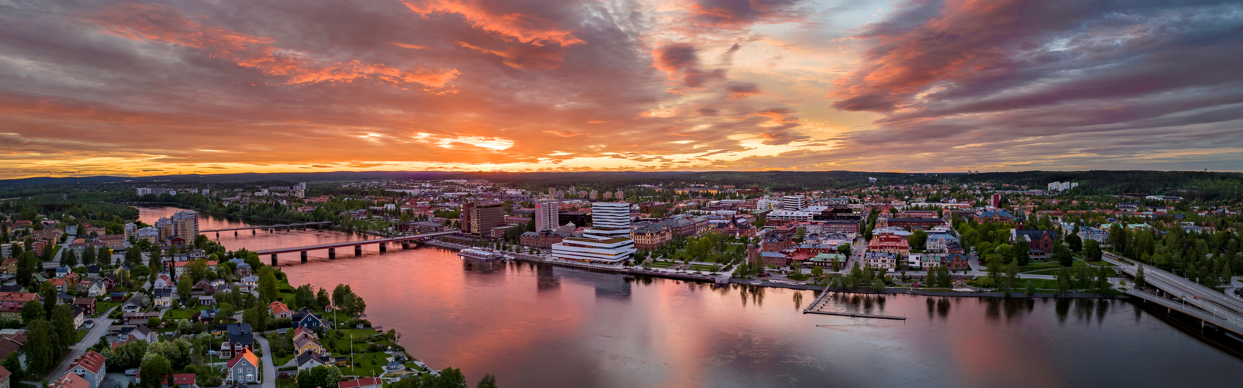Vy tagen med drönare från södra sidan av Umeåälven mot Umeås centrum med kulturhuset Väven i fokus. Himlen är röd-orange i solnedgången.