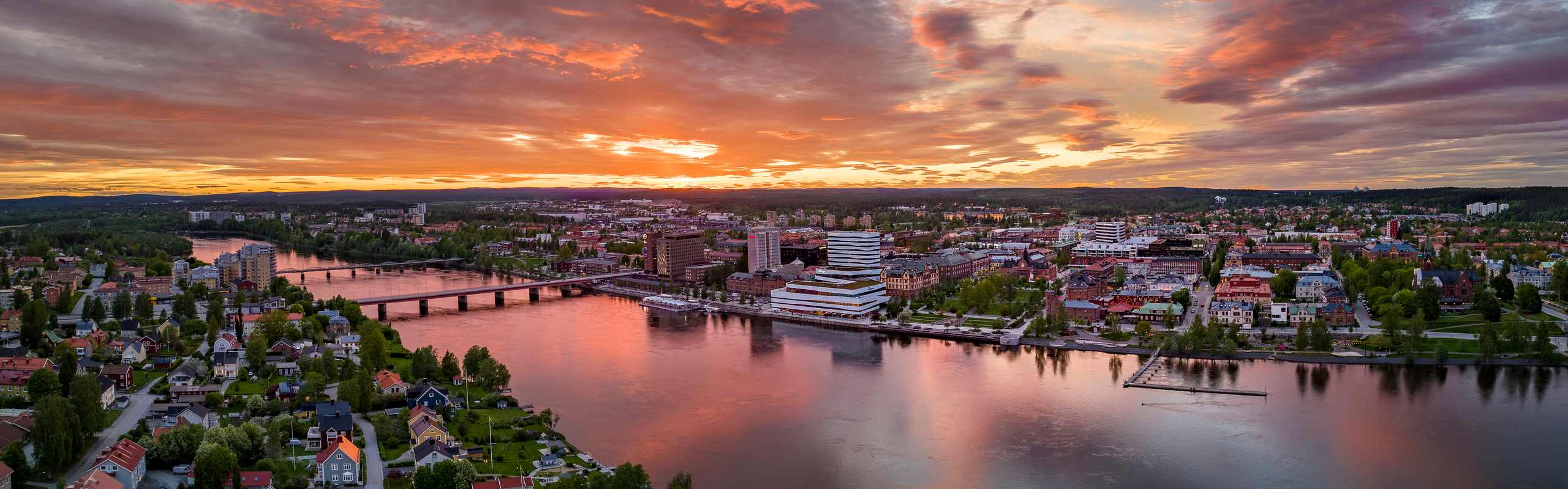 Vy tagen med drönare från södra sidan av Umeåälven mot Umeås centrum med kulturhuset Väven i fokus. Himlen är röd-orange i solnedgången.
