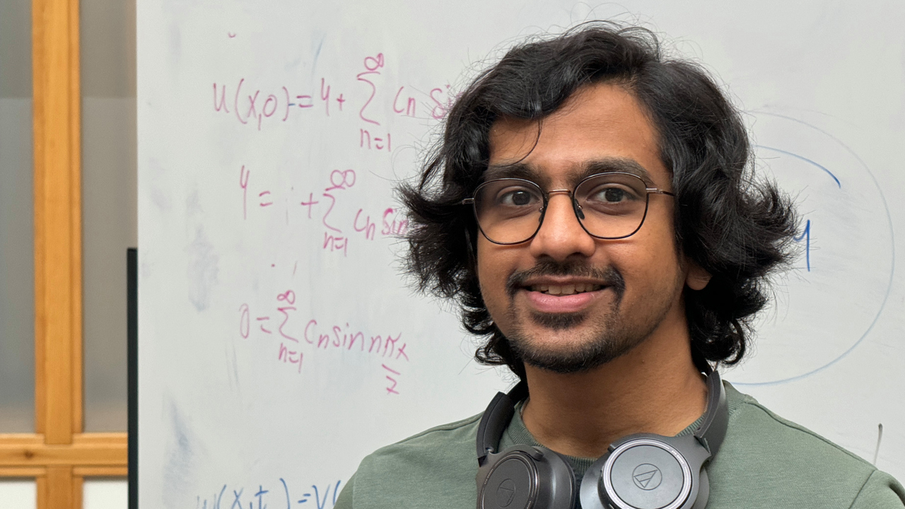 Hemamshu står framför en whiteboard med matematiska beräkningar. Han bär grön tröja och glasögon och har hörlurar runt halsen.