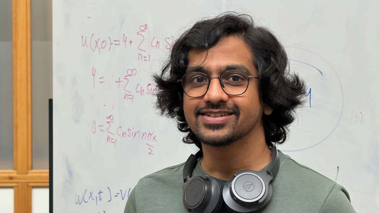 Hemamshu står framför en whiteboard med matematiska beräkningar. Han bär grön tröja och glasögon och har hörlurar runt halsen.