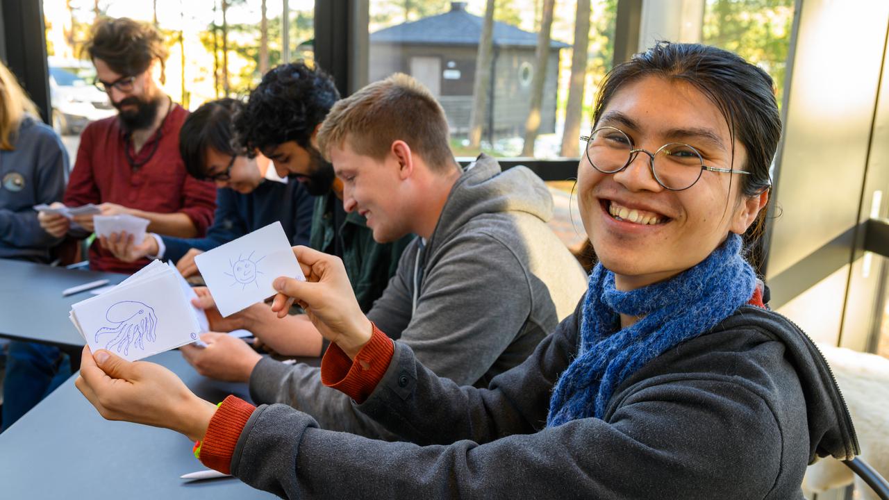 A group of people sitting around a table. A person with glasses smiles and shows two drawings, one of an octopus and the other of a sun.