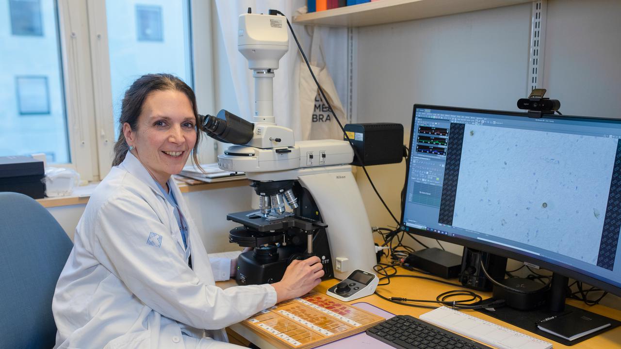 Karin Forsberg sitter vid ett mikroskop på sitt arbetsrum och tittar glatt mot kameran.