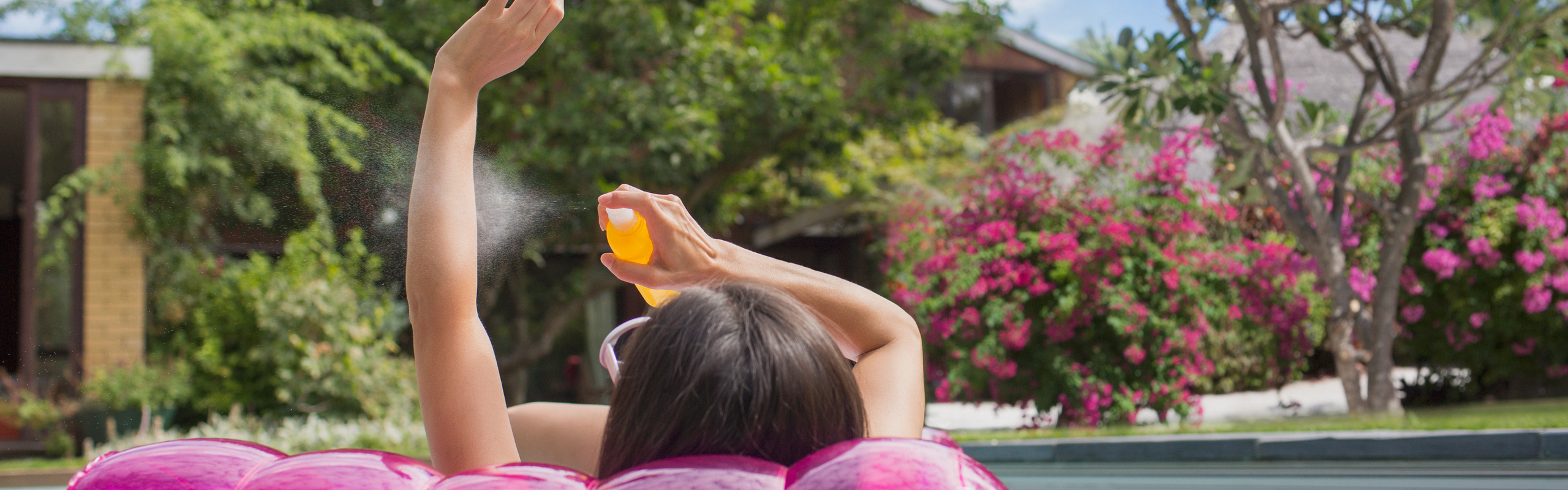 En person ligger på en rosa uppblåsbar madrass i en pool och sprutar solskyddskräm på sin arm. Gröna träd i bakgrunden.