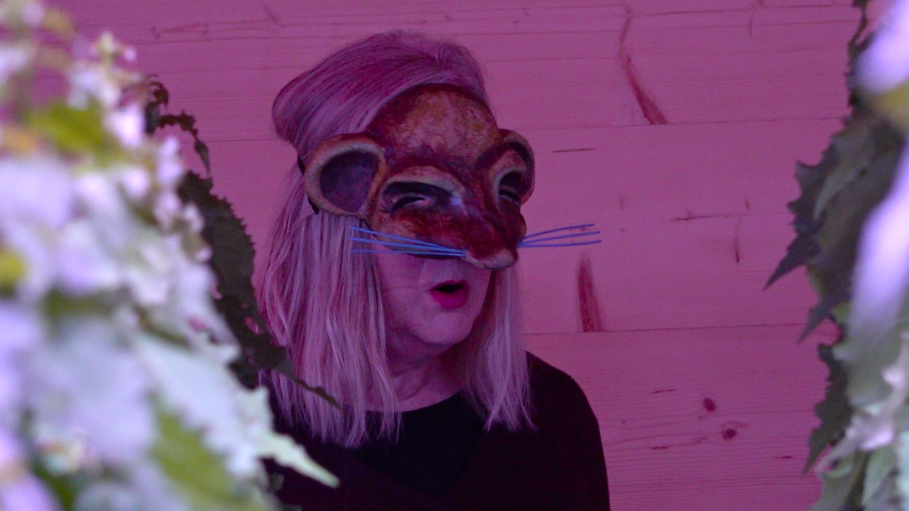 A blonde woman with a black t-shirt a face mask looking like a mouse, sits on a stage with pink light, and some hand made trees in the foreground.