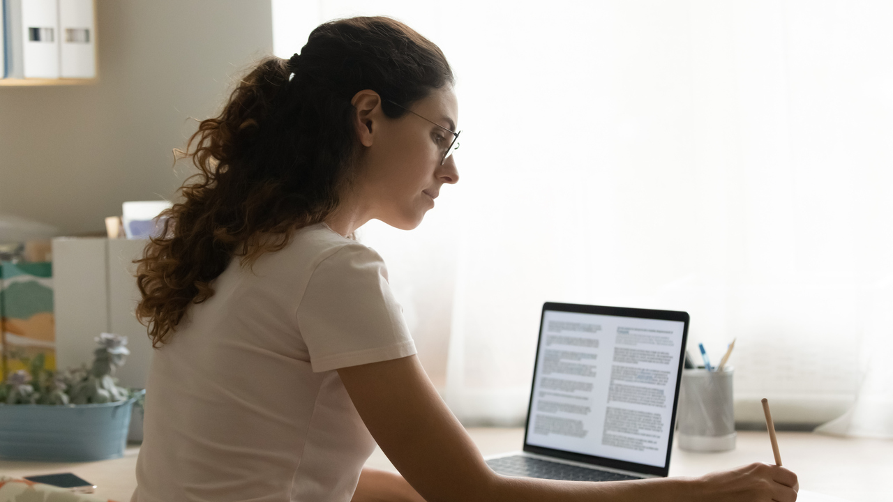 En kvinna sitter vid ett bord med laptop och headset, böcker och antecknar i ett block.