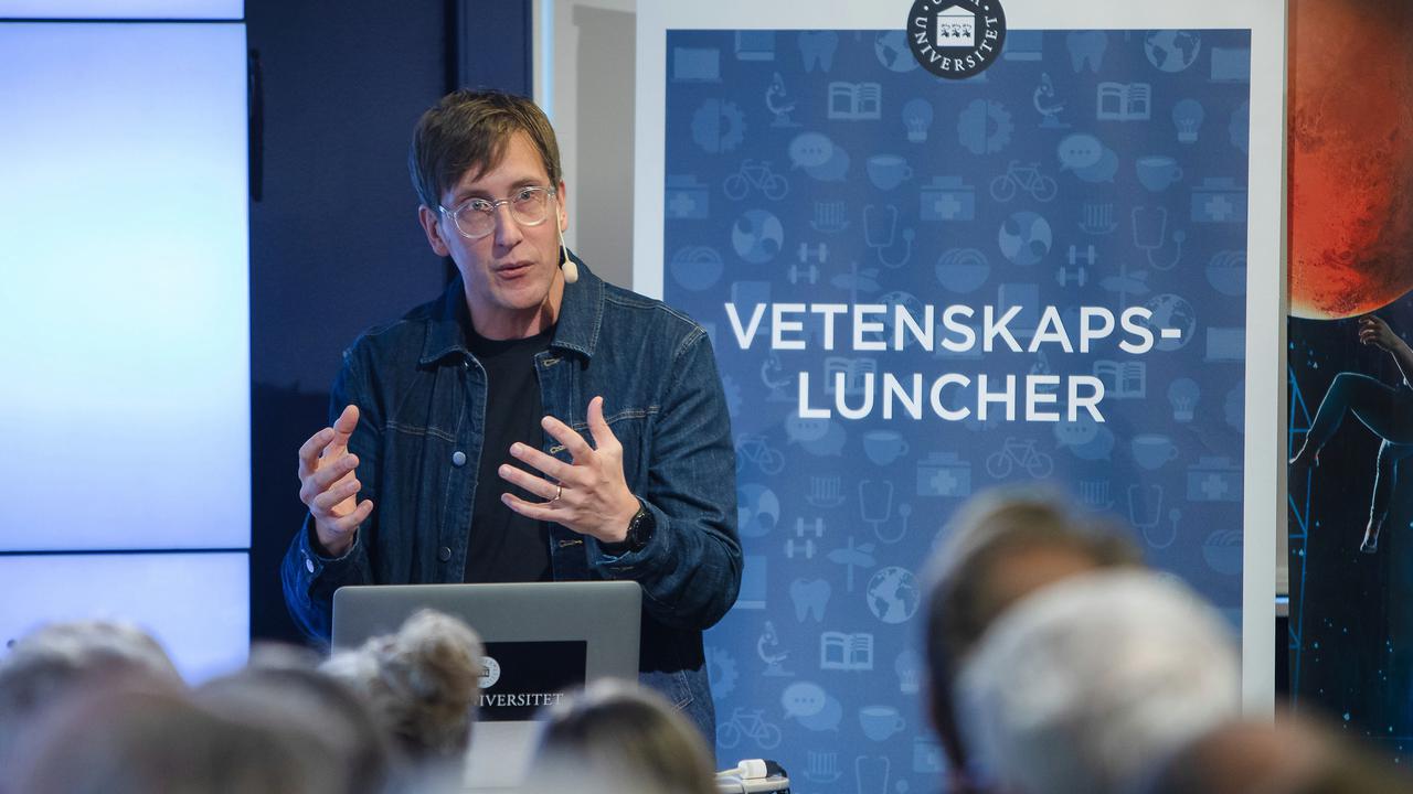 Föreläsare pratar inför publik, rollup med ordet vetenskapsluncher syns i bakgrunden.