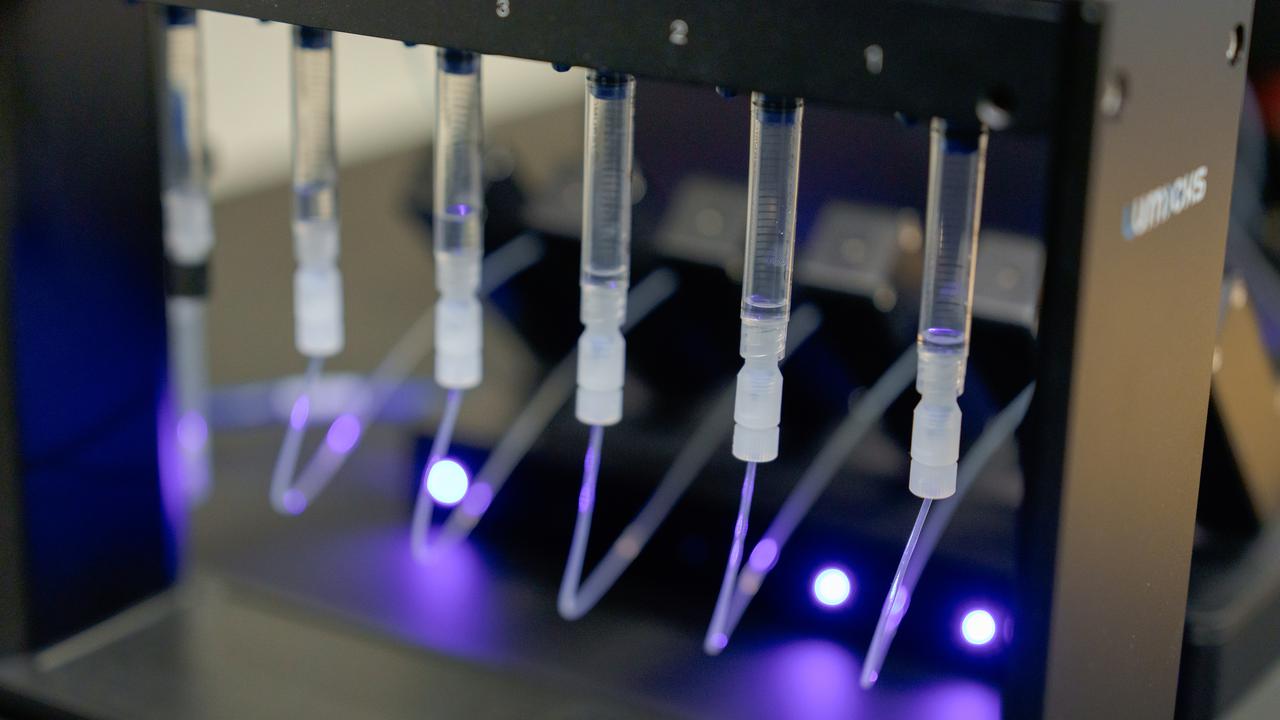 Sex transparenta kapillärer som hänger i ett svart stativ är anslutna via plastpluggar till små transparenta rör som belyses av ett violett ljus.