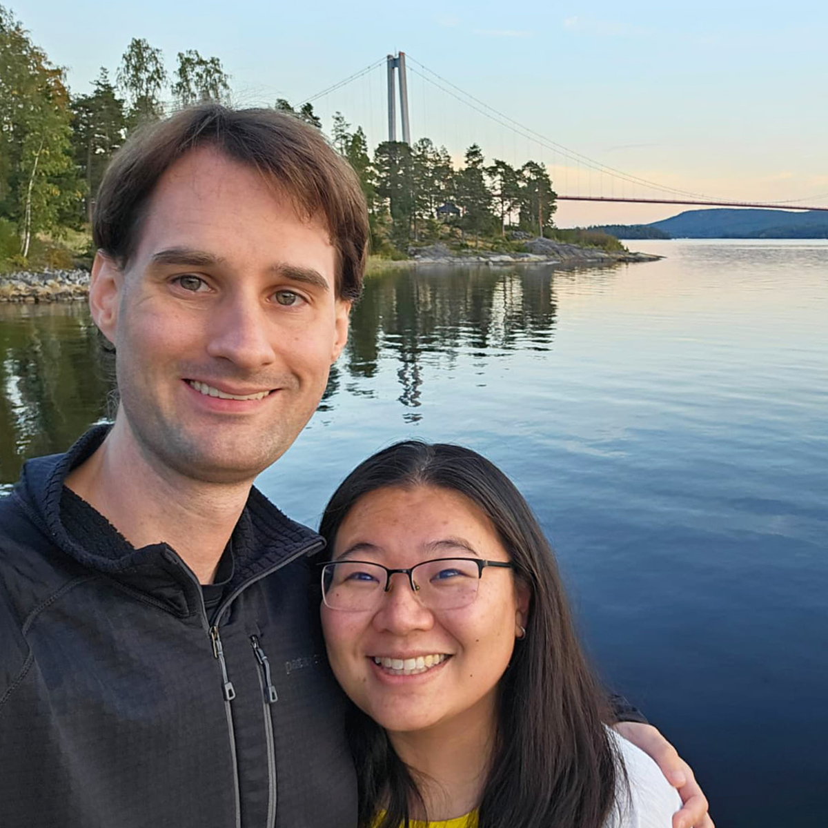 Par som tar selfie framför Höga kusten-bron i solnedgång.