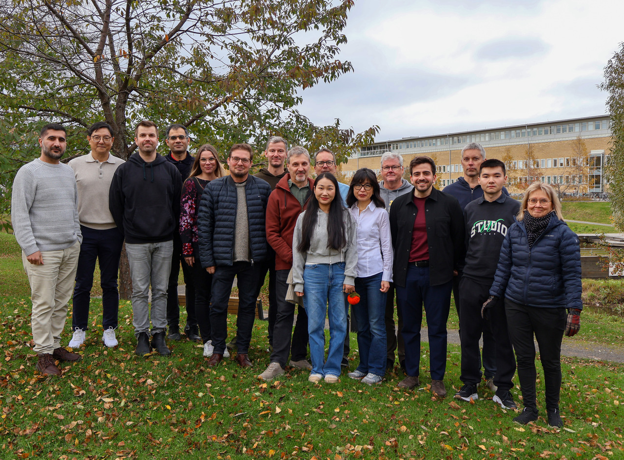 En grupp på sexton personer står utomhus på en gräsmatta med höstlöv, framför träd och ett universitetsbyggnad i bakgrunden. Personerna står i två rader och ler mot kameran.