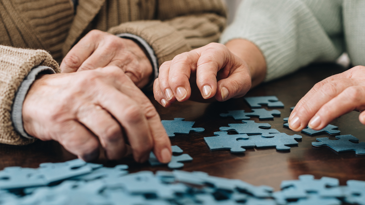 Två äldre personer lägger pussel tillsammans. Närbild på händer.