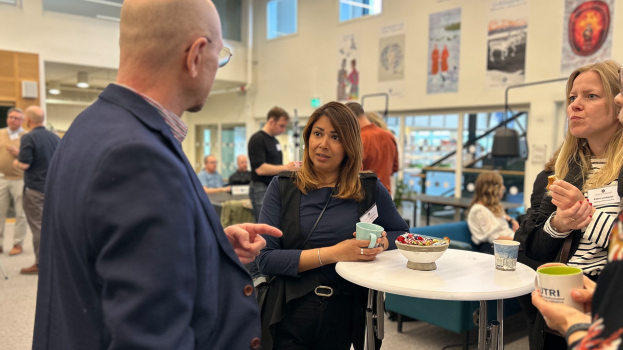 Fyra personer står samlade kring ett bord och pratar med människor minglande i bakgrunden.