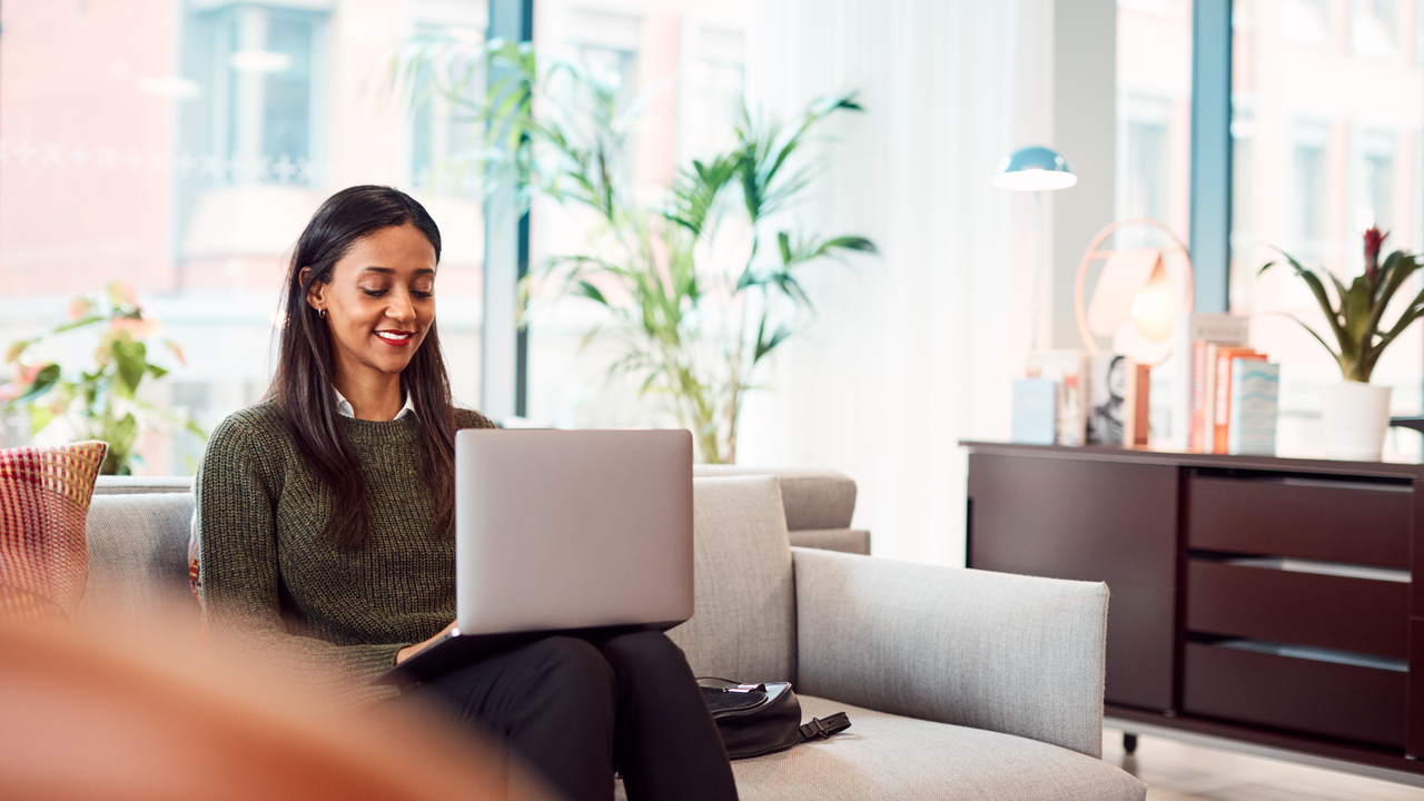 Glad kvinna sitter med laptop i beige soffa i kontorsmiljö.