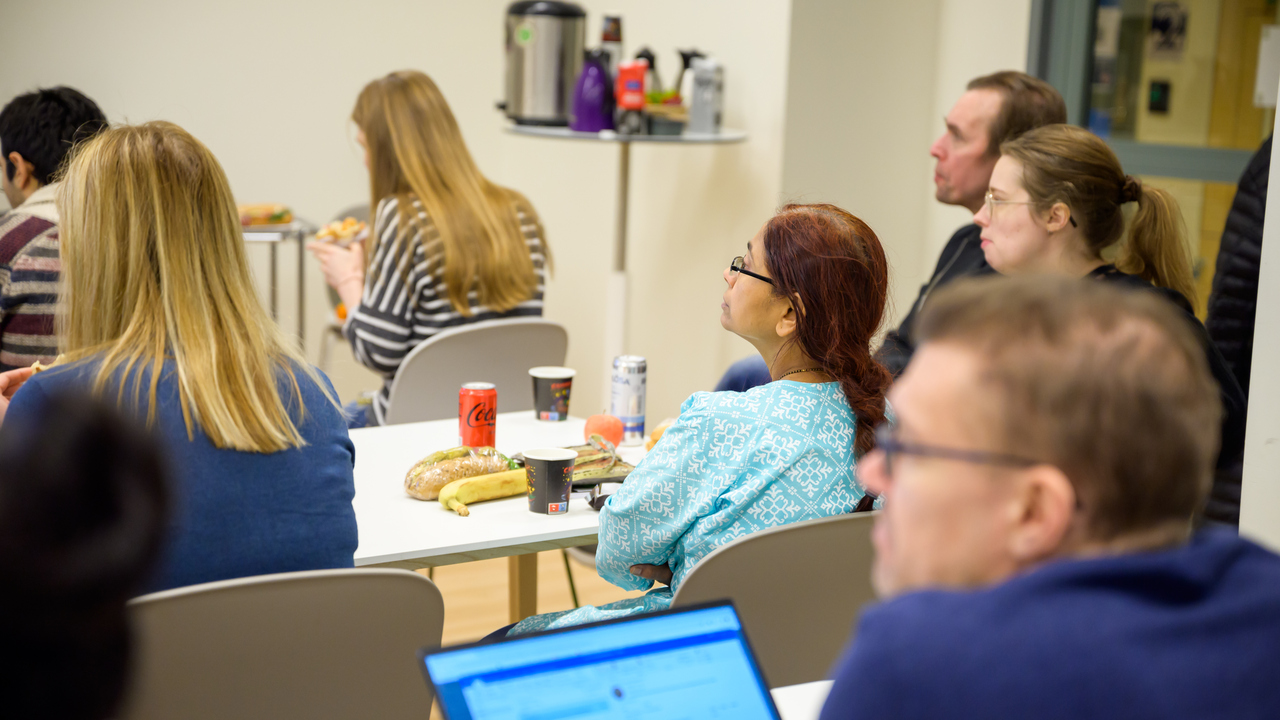 Six researchers are seated and looking forward towards a speaker who is not pictured. There are different items in the photo, like an open laptop, and lunch items.