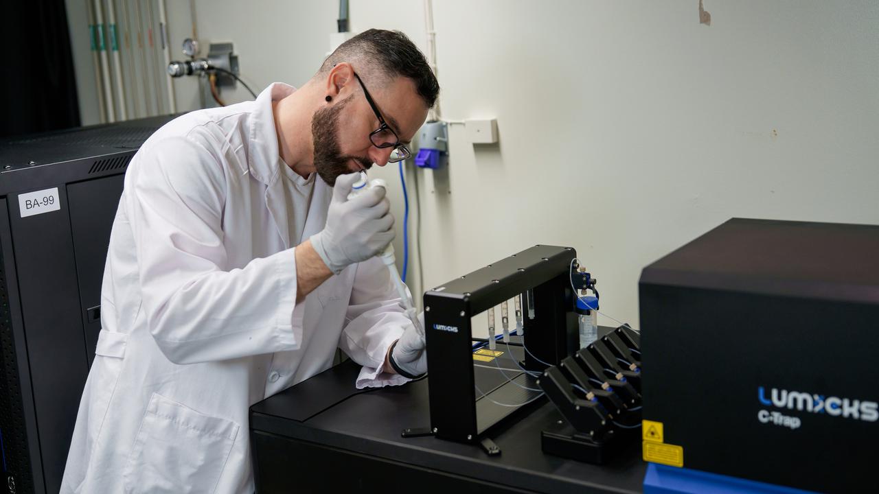 Rubén Casanova Sáez wearing a white labcoat and gloves is holding a small device in his hands that is connected to small pipes.