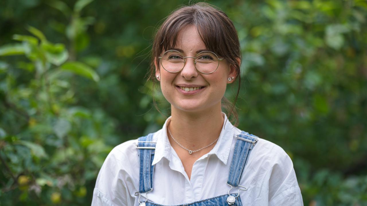 En kvinna med glasögon och overall står framför träd