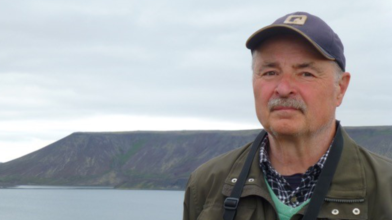 Bild på Lennart Bondesson som utomhus med en sjö och berg i bakgrunden. Han bär en keps och en grön jacka och ser direkt in i kameran.