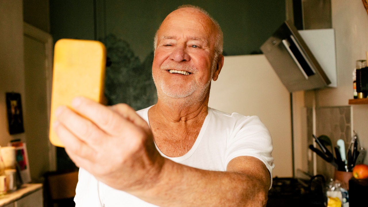En glad äldre flintskallig man med lätt skäggstubb och vit tröja står i kök och tittar i sin mobiltelefon