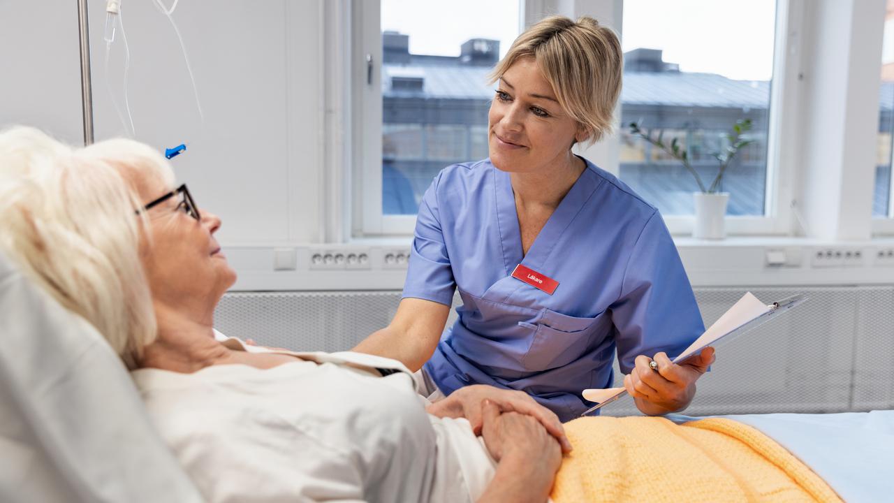 Sköterska sitter bredvid en säng där en äldre kvinna ligger