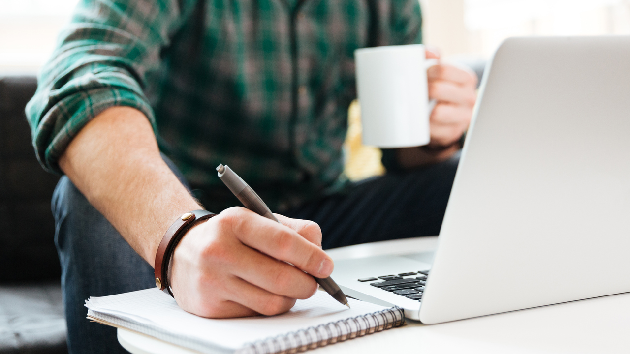 Person sitter i en soffa och arbetar. Har dator, kaffekopp och block där han skriver.